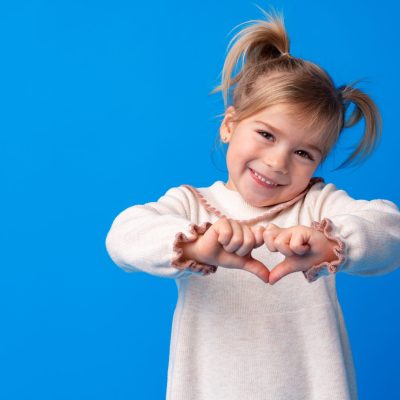 Happy little girl making heart shape by hands on blue background, close up